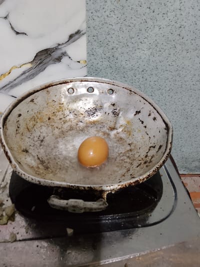A single, brown egg is positioned in the center of a weathered, metal frying pan, which rests on a gas stovetop. The pan appears to be slightly tilted, and water or oil is visible around the egg. The background features a textured, light blue wall and a section of a marbled backsplash. The overall scene suggests food preparation in a domestic setting, possibly in Merbopuntong, Indonesia.