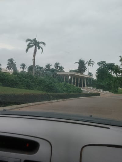 The image, captured from a vehicle's interior, shows a view of a building with a colonnaded entrance and a set of stairs leading up to it. Tall palm trees and lush green vegetation surround the structure. The sky is overcast. The building is located in Iju, Nigeria, near a paved road. The foreground reveals part of a car's dashboard.