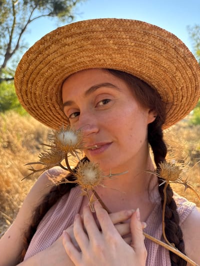 A close-up, waist-up portrait of a woman in a straw hat stands outdoors in a field of dry, golden grass under a clear blue sky. The woman has long, dark brown braided hair and is wearing a pink textured top. She is holding a dried thistle plant with several seed heads near her face. Her hands are clasped in front of her chest. The background is softly blurred, with trees visible on the left and right. The scene appears to be in Katzrin, Israel.