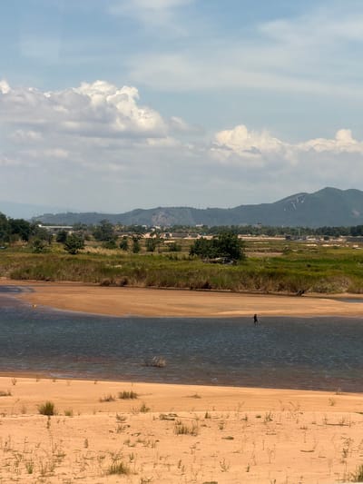 The image depicts a natural landscape near Tuy Hoa, Vietnam, characterized by a body of water, sandy shores, and vegetation. In the middle ground, a single individual is observed standing in the shallow water, appearing to be engaged in an activity. The water is bordered by a sandy beach that transitions into a landscape of grasses and scattered trees. In the background, hills and mountains rise, with some evidence of human infrastructure and construction visible on their slopes. The sky is partly cloudy, with a mix of white cumulus clouds against a light blue backdrop. The overall scene suggests a tranquil, somewhat undeveloped area.