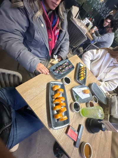 A group of people are seated around a wooden table laden with sushi dishes. Plates with various sushi rolls, dipping sauces, and a drink are visible. One person wears a gray puffer jacket and jeans. Another wears a white sweater. In the background, other patrons are visible through a glass wall. The table also holds a black object, a packet labeled "TEREA," and a long gray object.