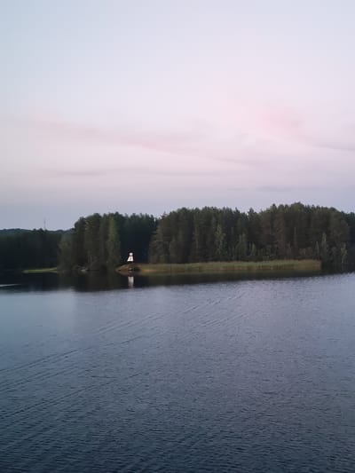 The image shows a body of water with a tree-lined shore in the background. A small white structure, possibly a lighthouse or marker, stands on the edge of the land. The sky above is a soft gradient of pale pink and blue, suggesting either sunrise or sunset. The water is dark and reflects the sky and trees. There are no people or animals visible in the frame. The overall scene is tranquil and natural. The location is identified as Nikol'skaya Gora, Russia.