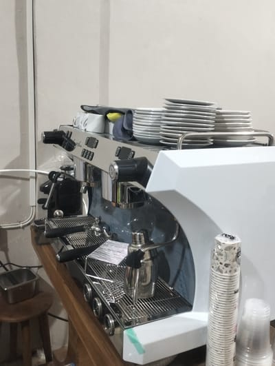 In Tingkeummanyang, Indonesia, a commercial espresso machine is centrally positioned on a wooden counter, its stainless steel and white body gleaming. Several stacks of white ceramic plates are arranged on top of the machine, and to its left, a stack of white ceramic mugs is visible. A stainless steel milk frothing pitcher rests on a rack in front of the machine, alongside other stainless steel accessories. To the right of the espresso machine, two stacks of disposable paper cups are placed. In the background, a white wall and a portion of a light-colored ceiling are visible. A small wooden stool with a metal container is situated to the left of the counter. The overall environment appears to be a cafe or a coffee preparation area.