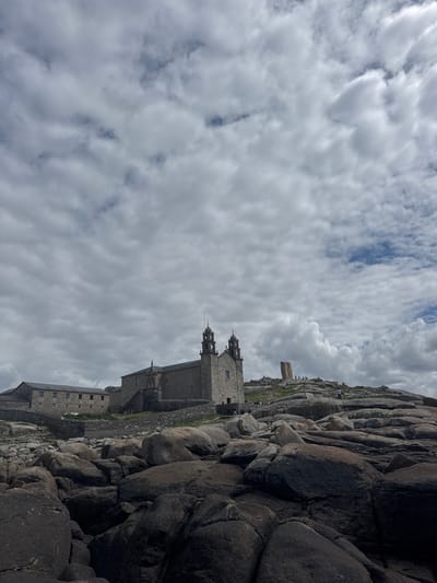 The image shows a view of the Sanctuary of the Virgin of the Barca in Muxía, Spain, located at coordinates 43.113, -9.221. The sanctuary, a stone building with a bell tower, is situated on a rocky outcrop. Several people can be seen walking around the grounds. The foreground consists of a large number of large, grey, moss-covered rocks that appear to have been smoothed by water.  The sky is cloudy with a mix of white and dark grey clouds.  The horizon is visible in the distance.  There is a smaller, more distant building, possibly a lighthouse, on a similar rocky outcrop to the right of the main sanctuary.