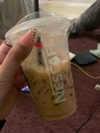 A person's hand, with light pink, almond-shaped nails decorated with black polka dots, holds a clear plastic cup filled with iced coffee. The cup has condensation on the outside and the Nescafé logo is visible on the side in white lettering. A black straw with a red accent is also present. The background shows a blurry, dark red carpeted surface and what appears to be a person's leg in dark pants. The scene suggests an indoor setting, possibly a living room or lounge area. There is no information in the image that indicates a specific location in Jajag, Indonesia.