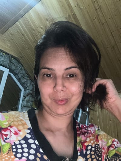 A woman is taking a selfie in what appears to be a room with a wooden ceiling and stone walls. The woman has dark brown hair and is wearing a colorful patterned shirt. She is looking at the camera and holding a strand of her hair with her right hand. The background includes part of a window and stone masonry. The lighting is dim, suggesting an indoor or evening setting.