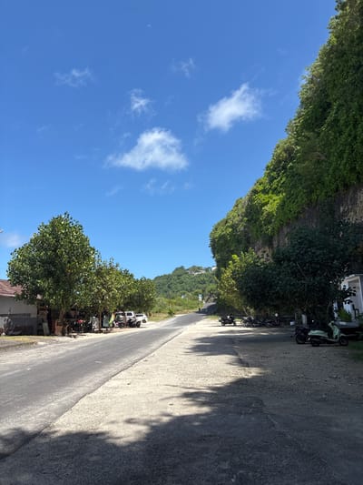 The image captures a road in Kutuh, Indonesia. The road runs diagonally across the frame, flanked on both sides by lush green vegetation and trees. On the right side of the road, several parked motorbikes are visible near a building, casting shadows on the road surface. The opposite side of the road also features dense vegetation. The sky above is a clear blue with scattered white clouds, and a sliver of the moon is visible in the upper left corner.