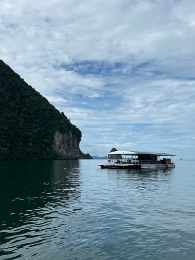 A long, narrow boat with a covered upper deck is floating on calm, dark water. The boat is painted white and dark blue, and it appears to have several people on board. There are railings around the deck, and some chairs and tables are visible. A white banner with some text in a foreign language is attached to the side of the boat.

To the left of the boat, a steep, lush green island rises from the water. The island is covered in dense trees and vegetation, with a rocky cliff face visible on its side. The sky above is partly cloudy, with streaks of white clouds against a light blue background. The sun appears to be shining, casting reflections on the water.

The scene suggests a leisurely boat trip or tour in a tropical or subtropical location. The overall mood is peaceful and serene. The location context provided places this scene in Khlong Khian, Thailand, known for its limestone karsts and emerald waters. The time of day is likely daytime, given the bright sky and reflections on the water. The weather appears to be pleasant, with a mix of sun and clouds.

Visible text: A white banner on the side of the boat has writing in a language that is not readily identifiable from the image.