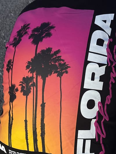 The image shows a piece of clothing with a design featuring the word "FLORIDA" in white block letters above a gradient sunset scene with black silhouette palm trees. The sunset colors transition from purple at the top to orange at the bottom. Black horizontal lines run across the sunset scene, representing a stylized beach horizon. Small text is visible on the lower right side of the garment. The location is not in Arinj, Armenia, since the image is of a piece of clothing related to Florida, USA.