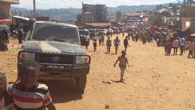 A dirt road, likely in a developing country, teems with people. A military-style Toyota truck occupies the left foreground, partially obscuring a young boy in a striped shirt. Behind the vehicle, people walk along the road. A second boy in a t-shirt is slightly off-center. In the background, buildings sit on hills, and a crowd stretches into the distance. The scene is busy and appears to be outdoors.