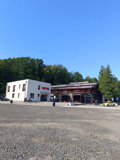 The image shows a motel and an adjacent open-air structure with a roof, located in Hațeg, Romania. The motel is a modern, white, two-story building with dark-framed windows. The word "MOTEL" is prominently displayed in red letters on its facade. In front of the motel, there is a gravel parking area. To the right of the motel is a larger, multi-story open-air structure with a complex wooden roof and supporting beams, likely a restaurant or covered seating area. This structure has large glass windows and an open ground floor with visible seating. Several people are present: two individuals walk near the entrance of the motel, and a few others are seen near the open-air structure. A yellow taxi with signage is parked on the road in front of the open-air structure. The background consists of a dense forest of green trees under a clear blue sky. The ground in the foreground is covered with gravel and scattered stones.