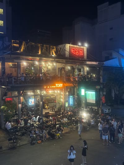 The image presents a vibrant nighttime urban scene, centered on a multi-story establishment identified by prominent illuminated signs reading "FACTORY" and "612 BISTRO BEER." This location is situated in Thuận Hóa Ward, Huế, Vietnam. The building features an open-air industrial aesthetic with exposed metal framework and multiple levels for patrons.
On the building's upper balcony level and the first-floor balcony, numerous individuals are seated at tables, engaged in socializing and dining or drinking under string lights and spotlights. The ground level of the establishment also hosts patrons seated at tables within a semi-open area.
Extending from the ground floor onto the asphalt street, a significant crowd of dozens of people, predominantly adults, are gathered. Many are seated at small tables and stools directly on the street, while others stand and interact. To the right of the ground floor entrance, a large white projection screen displays an image, likely a video or live event, which a segment of the street crowd is observing. A Vietnamese flag is mounted to the right of the projector screen. A white sedan is parked on the street further to the right. The entire scene is brightly illuminated by various artificial lights, contrasting with the dark night sky and dimly lit surrounding buildings in the background.