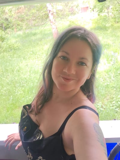 A woman is posing for a selfie. She has fair skin, blue eyes, and dark hair with blue and pink highlights. She is wearing a black dress with a floral pattern and black spaghetti straps. Her right arm is visible with a tattoo on her upper arm. She is standing on a white porch or balcony overlooking a green grassy area with trees. The background is blurred, suggesting depth of field. The location is possibly Свистуха (кузяевский С/о), Russia, although this cannot be confirmed from the image alone.