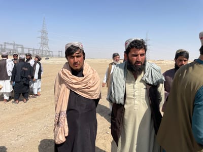 The image captures an outdoor scene in Pir Dust, Afghanistan, featuring approximately 10-12 adult males in an arid environment dominated by electrical infrastructure. In the foreground, two men are prominent. The man on the left, facing the camera, wears a black shalwar kameez, a patterned light beige shawl wrapped around his neck, and a light-colored embroidered cap. The man on the right, looking slightly left, has a full dark beard and wears a light beige shalwar kameez, a dark brown vest, and a light grey shawl draped over his shoulders, with a partially visible embroidered cap.
In the midground-left, a group of approximately 6-8 men, dressed in various traditional shalwar kameez, vests, and caps, are gathered and appear to be interacting. Another man is visible walking in the midground-right. The background features a large metal lattice transmission tower and multiple smaller steel structures, indicative of an electrical substation or power distribution point, with overhead power lines visible. A dark vehicle is partially visible among these structures. The ground is dry, light brown, unpaved earth with minimal vegetation, consistent with a desert landscape. The sky is clear blue, and the scene is brightly illuminated by direct sunlight.