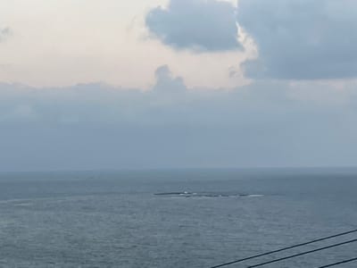 The image captures a wide view of the sea under an overcast sky. In the middle distance, a long, low-lying island or rock formation is visible in the water. Waves are breaking around the formation. The sky is filled with soft, muted clouds in shades of white and pale blue. The sea itself is a deep grey-blue, with subtle ripples on its surface. In the foreground, three dark, diagonal lines, likely power lines or cables, run across the bottom of the frame. The overall impression is one of a calm, serene, and somewhat melancholic seascape. The location of Mate Asher Regional Council in Israel is a coastal region in northern Israel, known for its scenic beauty and proximity to the Mediterranean Sea. This image is consistent with such a landscape.