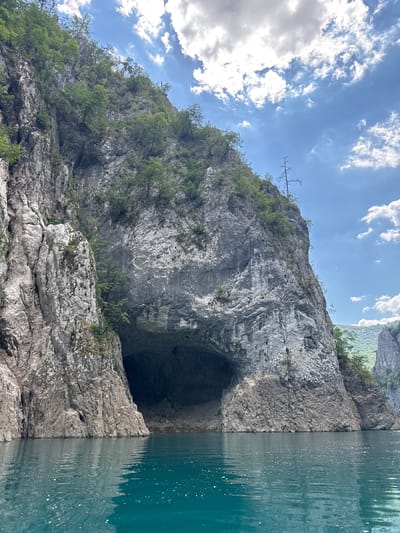 A wide-angle, eye-level shot captures a dramatic natural landscape with a large cave entrance carved into a steep, rocky cliff face. The cliff, textured with grey stone and sparse green vegetation, rises imposantly from the tranquil, turquoise water in the foreground. The water appears calm, with small ripples and white specks dotting its surface. Above the cliff, a bright blue sky is adorned with scattered, fluffy white clouds, suggesting a pleasant, sunny day. The scene depicts a remote, natural environment, likely a canyon or gorge, with the cave suggesting a potential geological feature of interest. There are no people or man-made structures visible in the frame. The overall impression is one of natural grandeur and serenity.