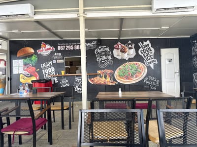 This media file captures an outdoor seating area of a restaurant, likely a grill and pizza place, with a distinct mural on the back wall. The mural features vibrant graphics of food items like a hamburger, pizza, and ice cream sundaes, alongside text related to the establishment. Visible text includes "TOP GRILL," a phone number "067 295 88," and various slogans like "ENJOY your FOOD," "Chilling AND Grilling," "STAY COOL EAT AN ICE CREAM," and "BBQ party." The word "TOALET" is also visible on a white door.

Several tables and chairs are set up, with some chairs featuring burgundy and yellow cushions. Two tables are visible in the foreground and middle ground, set with bottles of water and small containers. In the background, there's a glimpse of the restaurant's interior, including stacks of pizza boxes. Air conditioning units are mounted on the ceiling above the seating area, suggesting a warm climate. The scene appears to be during the daytime, under an overcast sky. There are no people visible in the image, creating a sense of quiet anticipation. The overall impression is that of a casual dining establishment ready to serve customers.