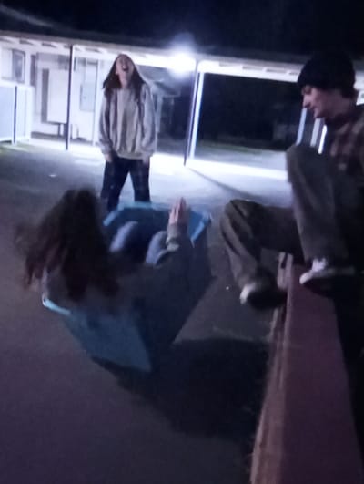 The image depicts a nighttime outdoor scene in Mirbozor, Uzbekistan, featuring three individuals engaged in a recreational activity. In the center-left foreground, a person with long dark hair is partially visible, positioned inside a light blue plastic container or bin lying on its side on a dark ground surface. Their hands are raised. In the left-center background, a female individual with long dark hair stands upright, wearing a light-colored sweatshirt and dark patterned pants. Her mouth is wide open, suggesting laughter or an exclamation. In the right foreground, a male individual wearing a dark beanie, a plaid shirt, and dark pants is seated on a reddish-brown ledge or wall, with his legs dangling over the side. He is looking towards the center of the scene, exhibiting a slight smile. The background consists of a single-story building structure with multiple openings and white pillars, illuminated by overhead artificial lights, casting shadows. The ground surface is dark.
