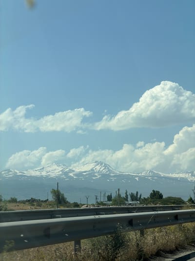 The image depicts a wide-angle view of a mountain range with snow-capped peaks under a clear blue sky dotted with fluffy white clouds. In the foreground, a grey metal guardrail runs horizontally across the lower portion of the frame, separating the road from the dry, scrubby vegetation. Behind the guardrail, a rural landscape unfolds with scattered trees, a few buildings, and power lines. The overall scene suggests a scenic vista, likely captured from a vehicle traveling along a road in Proshyan, Armenia, given the context. There are no people or animals visible in the frame. The lighting indicates it is daytime, likely mid-morning or afternoon, with the sun illuminating the scene.