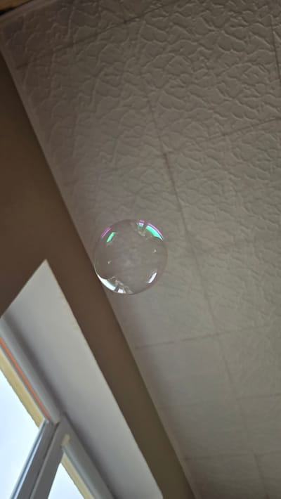 The image shows a single, transparent soap bubble with iridescent colors reflecting off its surface. The bubble is floating in the air, suspended against a textured ceiling. To the left of the bubble, a portion of a window frame and the sky outside are visible. The scene appears to be indoors, likely in a residential setting in Atkočiai, Lithuania, given the context. The ceiling has a patterned surface and faint grid lines, possibly indicating acoustic tiles. The overall lighting suggests it is daytime, with light coming from the window.