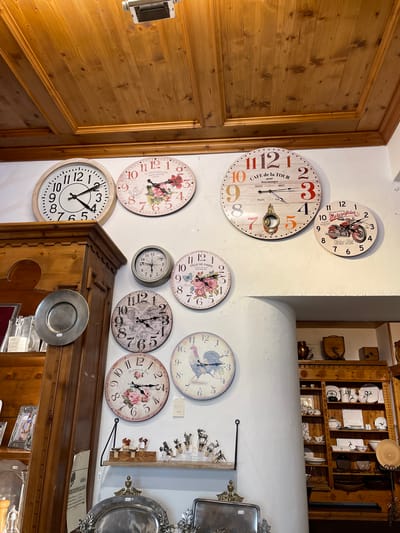 This media file shows a collection of decorative clocks displayed on a white wall, likely in a shop or home interior. The clocks feature various designs, including floral patterns, a motorcycle motif, and classic Roman numerals. Some of the clocks have visible text such as "CAFE de la TOUR", "VILLE DE PARIS", and "Motorcycles". To the left, a section of a wooden door is visible, indicating a rustic or traditional setting. To the right, a wooden shelving unit filled with plates, cups, and other decorative items is partially visible, suggesting a gift shop or a curated display. A metallic charger plate is hanging on the front of a wooden cabinet below some of the clocks. The overall impression is one of charming, vintage-inspired decor. The lighting suggests it is daytime, and there are no indicators of the weather. The location context suggests this might be a shop in Cortina d'Ampezzo, Italy, known for its alpine charm and artisan shops.