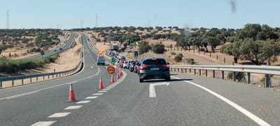 The image depicts a traffic jam on a highway in a rural, arid landscape with scattered trees. The day appears to be sunny and clear, suggesting warm weather. The road is paved, with multiple lanes separated by white lines. Traffic cones and a speed limit sign showing "40" are visible, indicating road work or some form of traffic control. A van with flashing lights and an orange warning sign on top is positioned at the front of the stalled traffic, likely related to the reason for the slowdown. Several cars are lined up behind it. In the distance, power lines stretch across the horizon, and the landscape is dotted with more trees. The overall scene suggests a frustrating delay for motorists. The license plate on the dark grey car in the foreground reads "B078 LVZ."