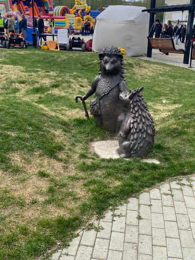 A bronze statue of two hedgehogs is on a patch of grass next to a paved area. One hedgehog is larger and has a cane, while the other is smaller and sitting on its back. In the background, there is a white tent and an inflatable play area, along with other people and objects that suggest a public event or gathering. This is located in the city of Kirovgrad, Russia.