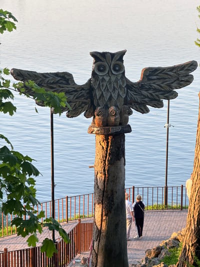 A wooden owl sculpture with spread wings, adorned with smaller owl carvings on the trunk, stands on a bank of the Kama River in the city of Krasny Klyuch, Russia. The sculpture is positioned near a sidewalk with a metal railing where two people are visible: one is wearing a white shirt and white pants, the other is wearing a black jacket. A security camera is installed above the sculpture. There are trees visible on either side of the image.