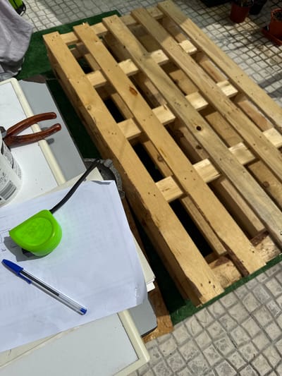 The image depicts an outdoor scene, likely a patio or garden area, with a wooden pallet as the central subject. The pallet is composed of light-colored wooden planks secured by nails. To the left of the pallet, a white surface is visible, possibly a table or workbench, on which a pair of pliers with red handles and a green tape measure are placed. A sheet of paper with some writing and a blue ballpoint pen lie on the white surface, near the tape measure. A partially visible grey fabric object is also present on the left. The ground beneath and around the pallet appears to be a combination of green artificial turf and patterned concrete tiles. In the background, several terracotta pots containing plants can be seen. The overall lighting suggests it is daytime. The location is specified as Almada, Portugal.