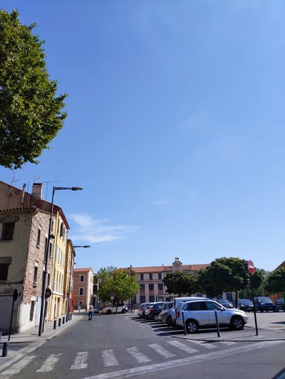 The image depicts a street scene in Perpignan, France, on a clear, sunny day. The perspective is from a street level, looking towards a street intersection with parked cars and buildings lining the sides. A prominent tree with green foliage is visible in the upper left quadrant.
On the left side of the street, aged buildings with brick and stucco facades are present. The building in the foreground on the left has a weathered appearance with visible brickwork and windows. A street lamp with a modern design is mounted on a pole on the left side of the street. A pedestrian crossing is marked on the asphalt road.
In the middle of the street, a person is walking, carrying a white bag. They are wearing casual clothing.
On the right side of the street, a parking area is visible with several cars parked. These include a silver SUV, a white van, and various other vehicles. A red and white circular "no entry" sign is posted on a pole in the parking area. Beyond the parked cars, more buildings and trees are visible. The sky is a clear, bright blue with a few wisps of white clouds. The overall impression is of a typical urban environment in a Mediterranean climate.