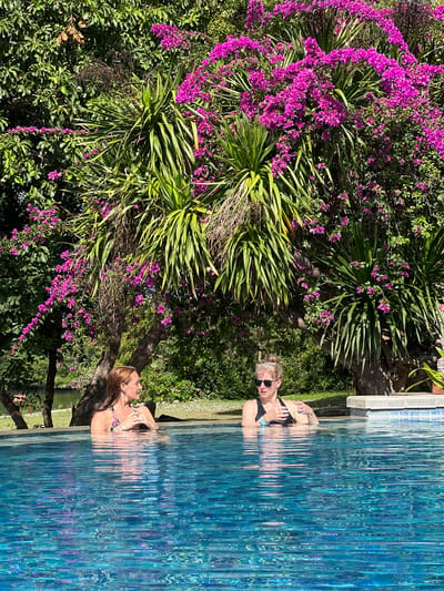Two women are in a swimming pool next to a lush garden with purple flowers. The woman closer to the viewer is lying back in the water, while the other woman is standing and facing forward. The location appears to be Gerokgak, Indonesia, given the tropical vegetation.