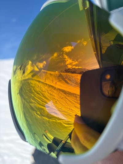The image displays a close-up of a white ski helmet equipped with a highly reflective, yellow-orange tinted goggle lens. The goggle's surface reflects a detailed winter scene consistent with the Zürs ski area in Austria. This reflection shows a clear blue sky with scattered white clouds, illuminating distant snow-capped mountain peaks. In the foreground of the reflection, a well-groomed ski slope is visible, exhibiting undulating snow textures and parallel ski tracks. Multiple dark figures, identified as skiers, are discernible descending the slope, with at least two red marker flags positioned along their path. On the right side of the goggle, a distorted reflection of a human hand, featuring a light pink fingernail, is visible, holding a dark-colored smartphone. The smartphone's multi-lens camera array is clearly reflected, indicating the device is actively being used to capture the image, presumably a selfie.