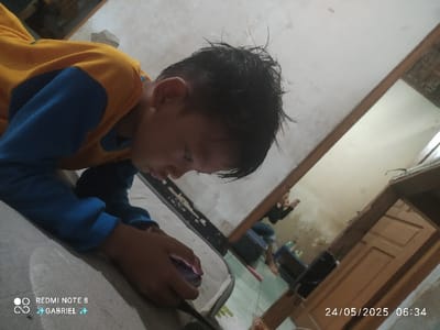 A young boy with short black hair, wearing a blue and yellow shirt, lies on a white matted floor, focusing intently on a smartphone held in his right hand. He is situated in an indoor environment, possibly a bedroom, with a mirror reflecting another person in the background. The reflection appears to be a woman in a blue shirt, holding a smartphone in her right hand and looking down.  The walls are a light beige color, and the floor is tiled. The time stamp on the image indicates that the photo was taken at 06:34 on 24/05/2025. The coordinates provided are for the city of Kebumen, Indonesia, but it's unclear if this is the exact location of the image.