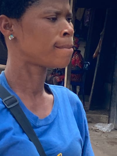 A young Black woman is shown in a medium close-up, from the shoulders up, in what appears to be a city setting. She is wearing a bright blue t-shirt and a black strap from a bag is visible across her chest. She has a small, square, iridescent earring in her left ear. Her head is turned to the right, and she is looking slightly downward and to the side, with her mouth partially open. The background is blurred and dark, with hints of fabric and structures suggesting an outdoor market or stall area. The overall lighting is dim, casting shadows and highlighting the contours of her face and neck. This scene is located in Nguzu Edda, Nigeria.