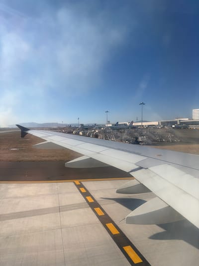 An airplane wing fills the right side of the frame, casting shadows on the concrete tarmac below, which is marked with intermittent yellow lines. In the background, an airport terminal area is visible under a clear blue sky. This includes parked aircraft, service vehicles, light poles, and a high-rise building in the distance. The perspective suggests the view is from inside a passenger aircraft window.