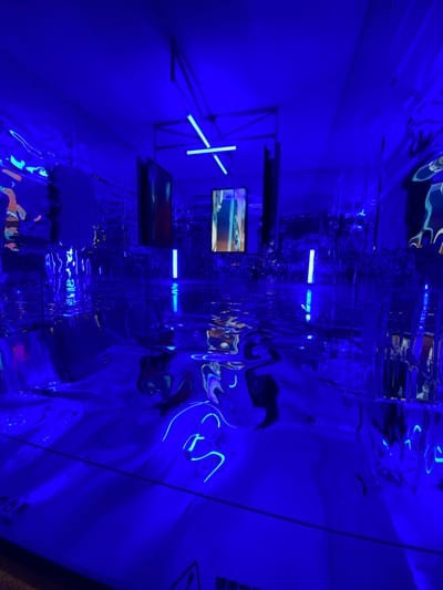 An art installation bathed in deep blue light features a reflective, wavy surface resembling water on the floor. Tall, thin blue light tubes are positioned vertically and horizontally, creating geometric patterns. Screens displaying abstract visuals are mounted on the walls and suspended from the ceiling. The walls themselves are covered in a crinkled, highly reflective material, mirroring the blue light and the artwork. The scene is dimly lit, with the dominant blue hue creating a surreal and immersive atmosphere. The location is specified as Jinniu District, China.