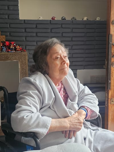 An elderly woman sits in a wheelchair, wearing a light gray robe over a pink polka-dot top. Her hands rest on her lap. She has gray hair and is looking upward. Behind her is a black brick-patterned wall adorned with figurines and a mirror. A wooden sliding door mechanism is visible above the wall. This scene is in Magdalena del Mar, Peru.