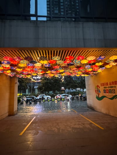 This image depicts the entrance to a building, possibly a mall or commercial establishment, in Longta, China, during twilight hours. The entrance is adorned with an overhead canopy of numerous colorful, decorative umbrellas or lanterns, creating a vibrant and festive atmosphere. The umbrellas are illuminated from within, casting a warm glow. Below the canopy, the ground is wet, indicating recent rain, with visible reflections on the pavement. Several vehicles, including cars and motorcycles, are parked in the vicinity, some with their headlights on. A few people are visible, some walking and others attending to the parked vehicles. The building facade features a large mural on the right wall, depicting a tree with colorful leaves and several icons below it, including what appears to be a recycling symbol and four square boxes with symbols inside. The overall scene suggests a bustling urban environment at the end of the day, with the decorative lighting adding an artistic touch to the entryway.