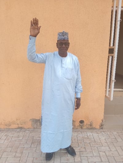 An adult male stands outdoors in Kabuga, Nigeria, centered against a plain, light orange-brown stucco wall. He wears black-rimmed glasses, a patterned cap, a light blue long-sleeved traditional garment, and dark slip-on shoes. A watch is visible on his left wrist. His right arm is raised vertically, with the hand open and palm facing forward. The subject has a slight smile. The ground is paved with light-colored interlocking bricks arranged in a herringbone pattern. A partial white door or window frame is visible to the right of the man against the wall, which exhibits dark discoloration near its base.