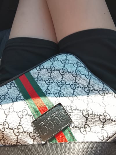 The image shows a close-up of a person's thighs and a handbag. The person is wearing black shorts. The handbag is a Gucci GG canvas bag with the signature red and green stripe. A black leather tag on the bag reads "GUCCI MADE IN ITALY". The lighting suggests it is daytime, and the scene appears to be indoors, possibly in a car, given the dark background and the positioning of the bag. There are no strong emotional cues visible. The provided location context of Kableshkovo, Bulgaria, does not offer any additional visual information about the scene itself.