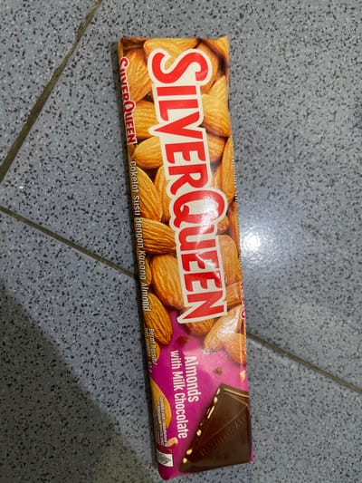 A Silver Queen chocolate bar with almonds and milk chocolate is lying flat on a gray, speckled tiled surface. The chocolate bar is oriented vertically with the top of the bar angled towards the top left of the image. The "SILVER QUEEN" logo is prominently displayed in large red letters on the front of the wrapper, which also features images of almonds. Text on the wrapper indicates "Almonds with Milk Chocolate" and details in Indonesian, such as "Cokelat Susu dengan Kacang Almond." The background is a close-up view of a tiled floor, appearing to be an indoor setting. No people or animals are visible in the image, and no specific city or location details are discernible from the visual information.