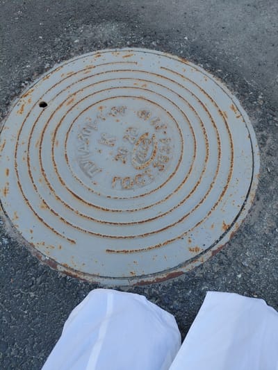 A slightly tilted, high-angle shot captures a circular, light grey metal manhole cover embedded in dark grey asphalt and gravel. The manhole cover is heavily worn, showing extensive reddish-brown rust along its concentric raised rings and overall surface, indicating oxidation. A small, dark circular hole is visible near the bottom-left edge of the cover.
On the right side of the frame, two white, folded objects, possibly fabric sheets or plastic bags, are partially visible, lying on the asphalt next to the manhole cover. The scene is static, illuminated by natural daylight, suggesting an outdoor setting.
Visible Cyrillic text on the manhole cover, typical of infrastructure in Russia (consistent with the Nadym context), includes:
* "ГОСТ 3634-99" (GOST 3634-99, referring to a Russian State Standard for manhole covers).
* "20 т" (20 tons), indicating a load capacity.
* "К" (K), likely an abbreviation for "канализация" (sewerage or drainage).
* "20.02", possibly a date or a specific code.
Additional, less legible text includes what appears to be "ТУ" (likely for "Технические Условия" - Technical Specifications) towards the bottom arc.