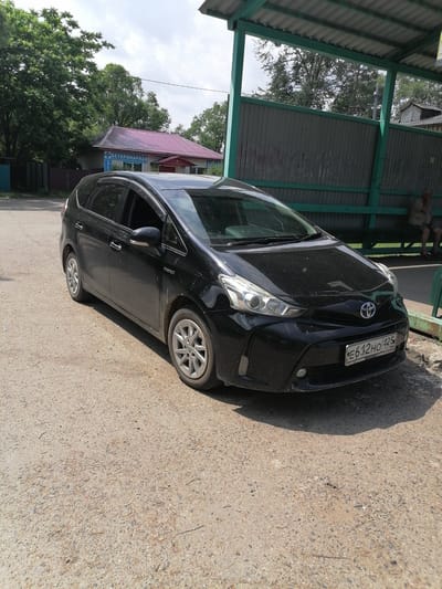 A black Toyota Prius is parked on a dirt road in front of a building with a red roof, partially covered by trees. The building has text on it that reads "ВЕТЕРИНАРНАЯ" which translates to "VETERINARY". To the right of the car, there is a green metal structure with a corrugated metal wall and a bench where a person is sitting. The license plate of the car reads "E612HO 425 RUS". The scene appears to be in the city of Novopokrovka, Russia.