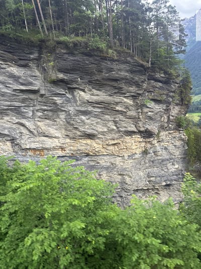 The image shows a rugged, stratified rock cliff face covered in layers of gray and tan rock. Dense green foliage and trees are visible at the top of the cliff and in the foreground. The background reveals a hazy mountain landscape with hints of green valleys and a cloudy sky. The scene appears to be a natural environment, possibly in a mountainous region of Switzerland, given the mention of Hüsen. There are no people or discernable actions depicted in the image.