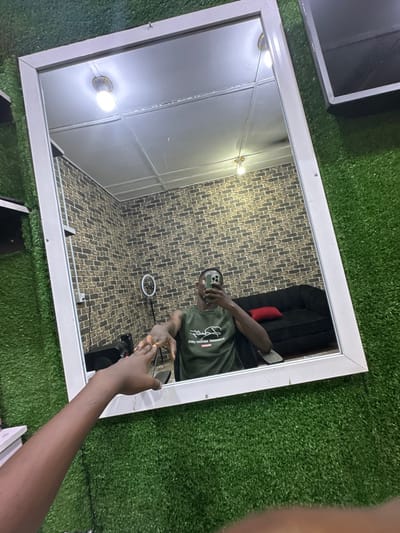 A mirror lies on a green artificial turf, reflecting a man taking a selfie. He's wearing a green shirt and holding a phone. The background features a brick-patterned wall, a black sofa with a red pillow, a ring light, and ceiling lights. Parts of objects, like a black box and a white stack, are visible near the mirror's edges. The mirror has a white frame.