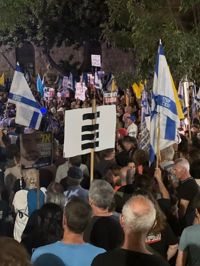 This nighttime image captures a dense crowd participating in a protest or rally in West Jerusalem, Israel, likely near Kikar Paris (Paris Square) or the Prime Minister's Residence, identifiable by the distinctive large, light-colored stone building and mature trees in the background. The scene is illuminated by artificial light sources.

Numerous individuals are visible, primarily adults, many seen from behind, facing forward in a common direction. People are holding a significant number of Israeli flags (blue and white with the Star of David). Several protest signs are prominently displayed. One large white rectangular sign features four thick black horizontal bars. Another sign depicts a man's portrait and bears Hebrew text that translates to "Don't leave us behind." Other signs further back contain Hebrew text such as "לא עוצרים 7.10" (Not stopping 7.10), potentially referencing the October 7th attacks, and "דו קיום" (Coexistence). The crowd is closely packed, indicating a large-scale gathering. No specific actions beyond holding signs and flags and standing are discernible, and no interactions between individuals are clearly depicted. The overall environment is urban, characterized by the historical stone architecture and mature trees, typical of central West Jerusalem.