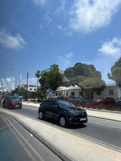 The image depicts a street scene in Paphos Municipality, Cyprus, under a clear blue sky with scattered white clouds, suggesting it is daytime. The perspective is from inside a vehicle, looking out.
The foreground features the edge of the road with double yellow lines, and a concrete curb. Several cars are parked or driving on the road. A black SUV is parked closest to the curb, with its rear facing the viewer. Further down the road, a red hatchback is visible.
Beyond the road, there is a sidewalk with some landscaping, including red flowers and trees. Buildings with white walls and balconies are visible in the background, along with a chain-link fence. A blue parking sign with a white "P" is mounted on a pole. The reflection of trees and the sky is visible on the glass, indicating the image was taken through a window.
The license plate on the black SUV reads "KOU-H21-5". The license plate on the red hatchback is partially visible and appears to be "TE2 713". The presence of palm trees and the architectural style of the buildings are consistent with a Mediterranean location. No people are clearly visible. The overall atmosphere is one of a sunny day in a moderately urban environment.
