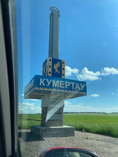 The image shows a monument marking the entrance to the city of Kumertau, Russia, located near the city of Ira. The monument is tall and white, with a decorative top. The words "КУМЕРТАУ" are written in large, silver letters on a blue sign. Below the blue sign, there is a smaller sign with the year "1953" inscribed on it. The monument is surrounded by gravel and a green field stretches out behind it under a bright blue sky with scattered white clouds. The photo appears to be taken from inside a vehicle, with part of the car's interior visible in the foreground.