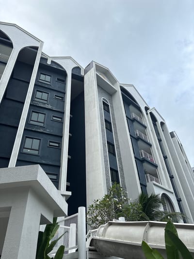 The image shows a low-angle view of a modern apartment building with a dark blue facade and white architectural accents. The building features a series of arched vertical elements and balconies with white railings. Several rectangular windows of varying sizes are visible on the dark blue sections. In the foreground, to the lower left, a white textured concrete structure is partially visible. To the right of this structure, lush green foliage and a large, white, curved object, possibly part of a water slide, are present. The sky above is overcast with a light gray hue. The overall scene suggests an outdoor setting, likely in a residential or resort area. The architectural style is contemporary, with a focus on clean lines and contrasting colors.