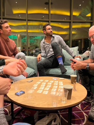 The image captures an indoor evening scene within what appears to be a hotel lounge, likely in Ein Bokek, Israel. Several individuals are gathered around a wooden table, engaged in a board or card game.
In the center, a man with a beard and glasses, wearing a checkered shirt and dark pants, sits on a light blue couch. He has a distinctive blue patterned sock on his right foot and is looking upwards and to his right. To his left, an older man with grey hair and glasses, in a grey shirt, is holding a cell phone, his smartwatch visible on his wrist.
In the foreground to the left, another man with short curly hair and a reddish-brown sweater is looking towards the bearded man, his hands clasped with another person's hand partially visible. Further to the right, a fourth person's arm, wearing a plaid shirt, is also visible near the table.
The focal point of the activity is a round wooden table. Laid out on the table are numerous small, rectangular game cards, although the text on them is illegible. There are also several clear plastic cups, two brown paper cups, and a blue board game box (possibly for a game like Codenames). The setup strongly suggests a social card or board game in progress.
The setting features large windows in the background, reflecting warm yellow artificial lighting from within the room, indicating it is nighttime. Beyond the windows, faint green elements, possibly foliage or outdoor decor, are visible. The floor is carpeted with a distinctive red and purple patterned design. The overall atmosphere appears relaxed and social, with participants engaged in the game.